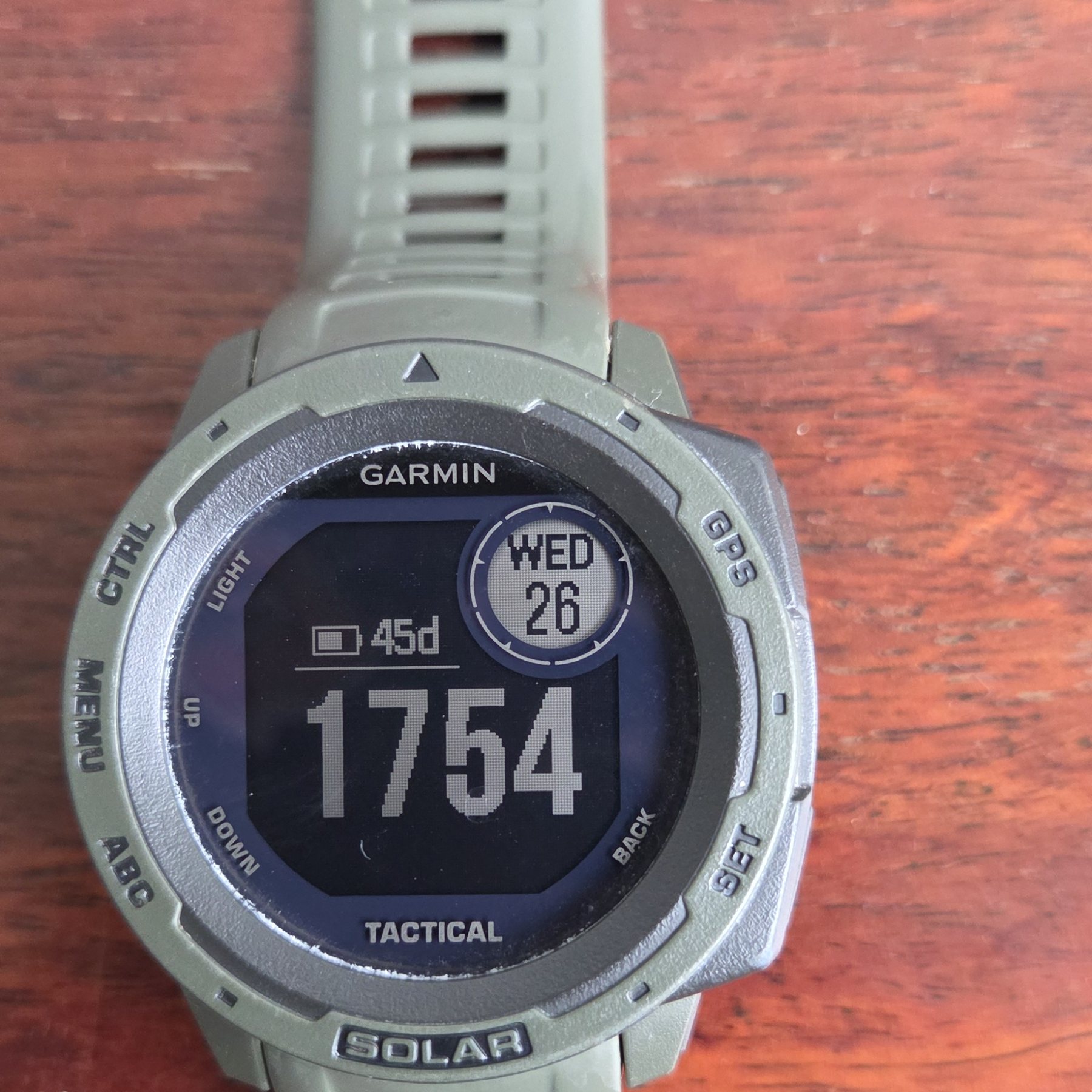 A Garmin Instinct Tactical with a sage green case and strap, its monochrome screen showing the time and a battery gauge.